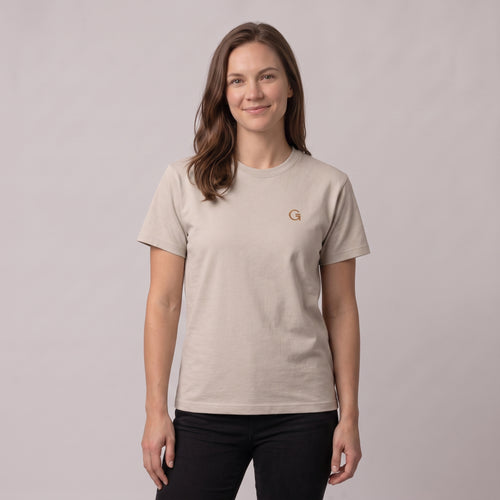 A woman modelling a sandstone t-shirt with a russet embroidered ‘Clobber & Gear’ logo on the left chest, paired against a light grey studio background.