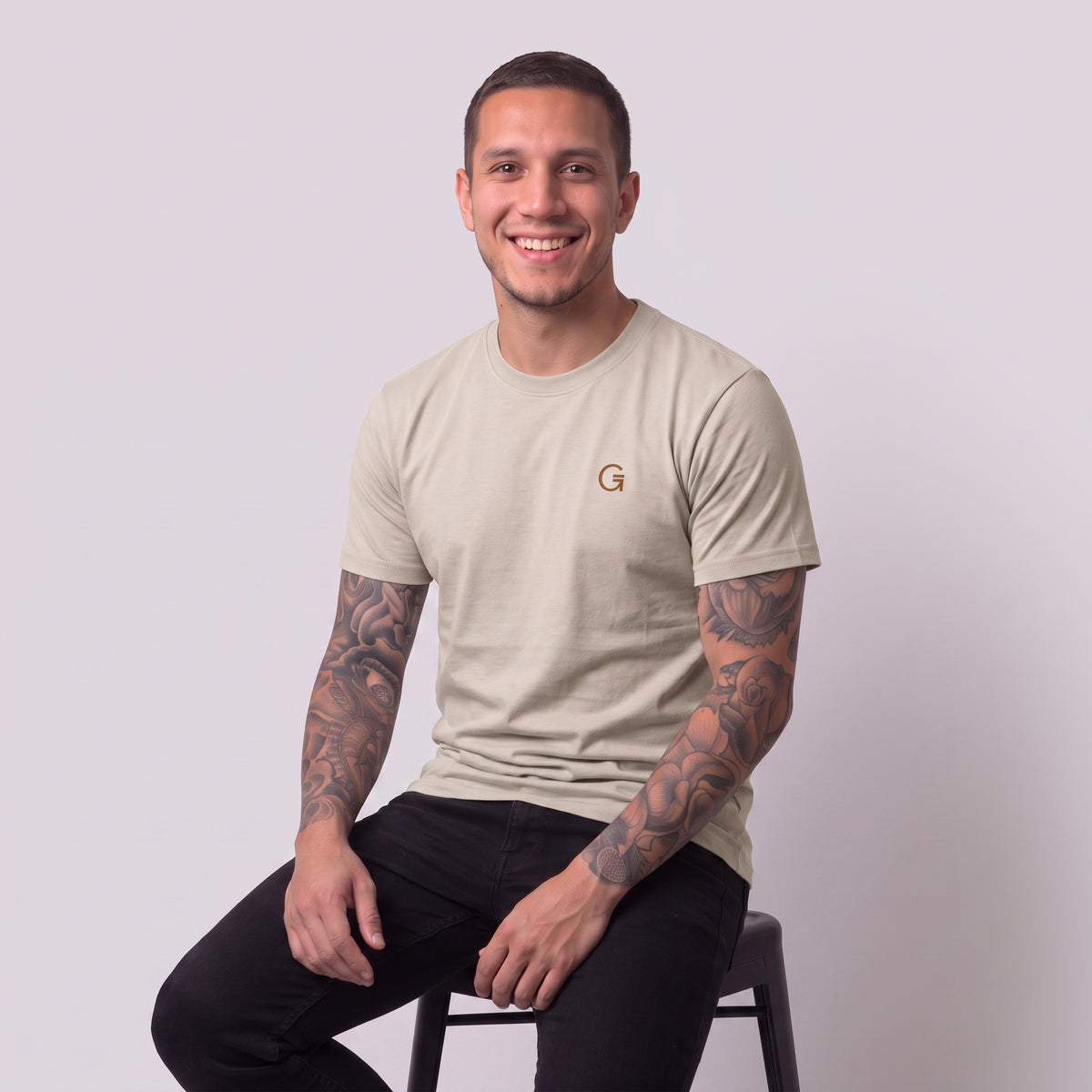A man modelling a sandstone t-shirt with a russet embroidered ‘Clobber & Gear’ logo on the left chest, paired against a light grey studio background.