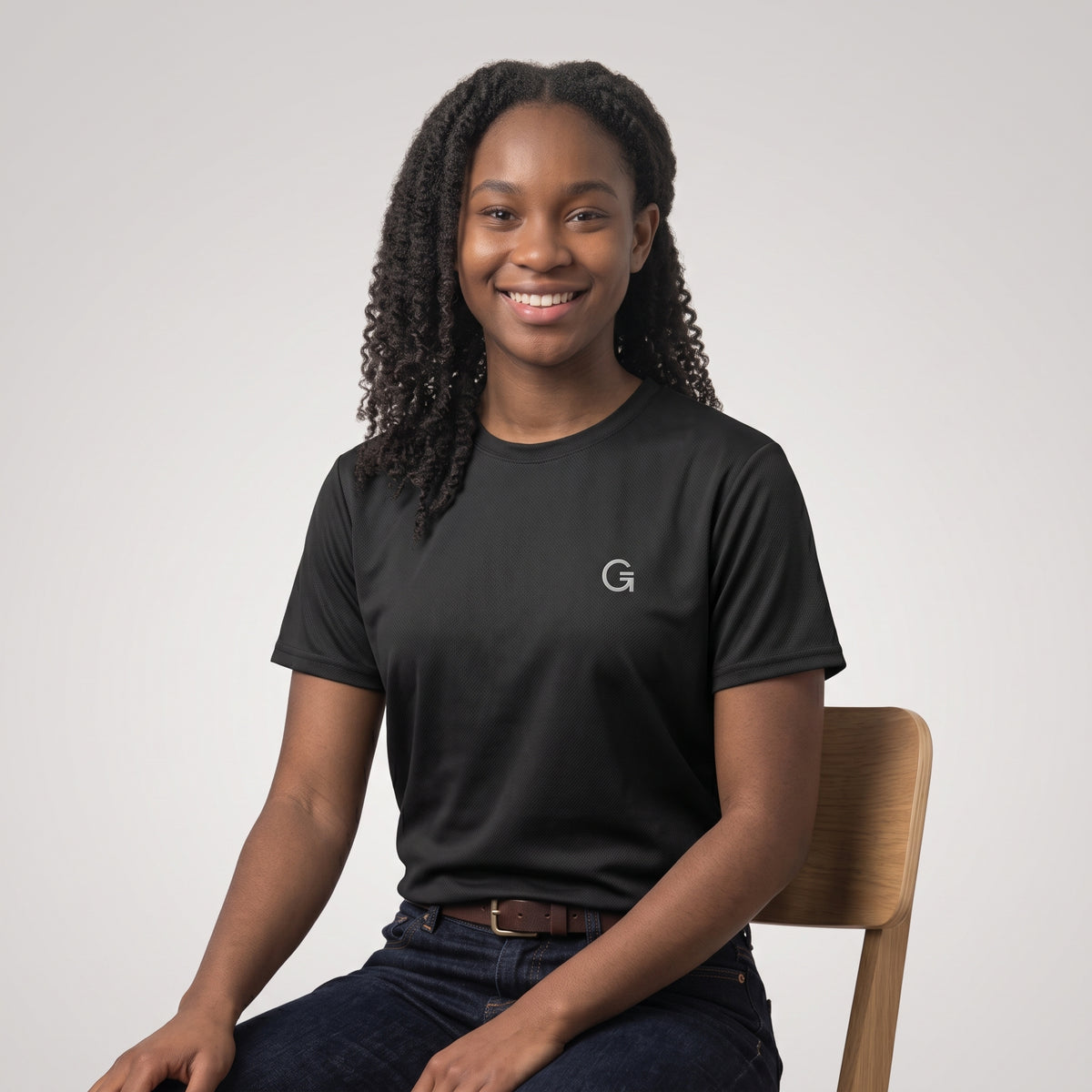 A woman wearing a black short-sleeve t-shirt with a white “Clobber & Gear” logo embroidered on the left chest, set against a light grey background.
