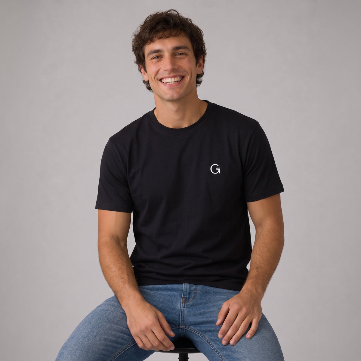 A man modelling a black tee with a white embroidered ‘Clobber & Gear’ logo on the left chest, paired against a light grey studio background.