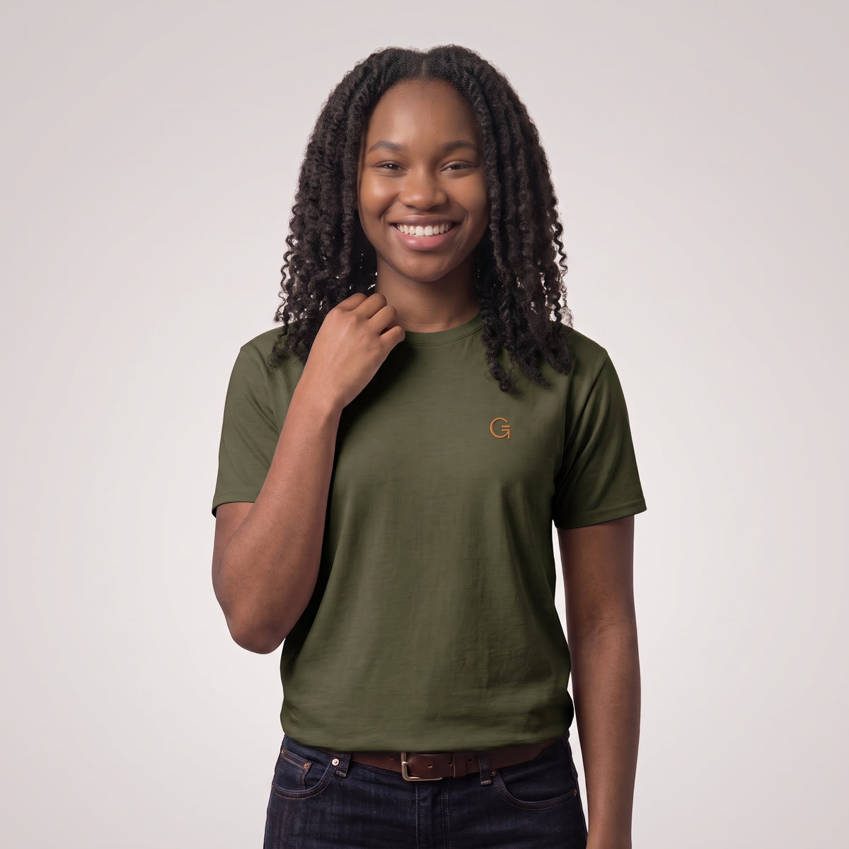 A woman modelling a military green tee with a burnt orange embroidered ‘Clobber & Gear’ logo on the left chest, paired against a light grey studio background.