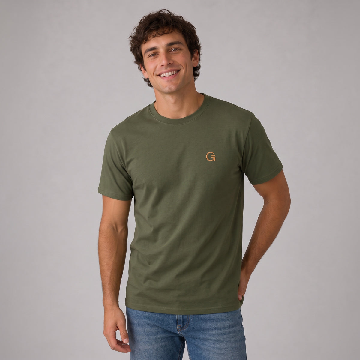 A man modelling a military green tee with a burnt orange embroidered ‘Clobber & Gear’ logo on the left chest, paired against a light grey studio background.
