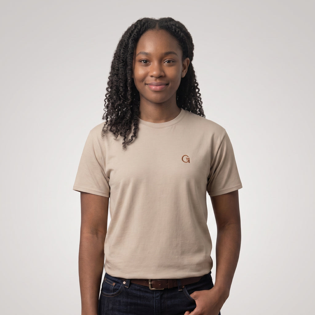 A woman wearing a sandstone short-sleeve t-shirt with a russet “Clobber & Gear” logo embroidered on the left chest, set against a light grey background.