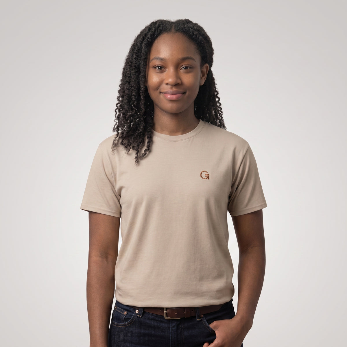 A woman wearing a sandstone short-sleeve t-shirt with a russet “Clobber & Gear” logo embroidered on the left chest, set against a light grey background.