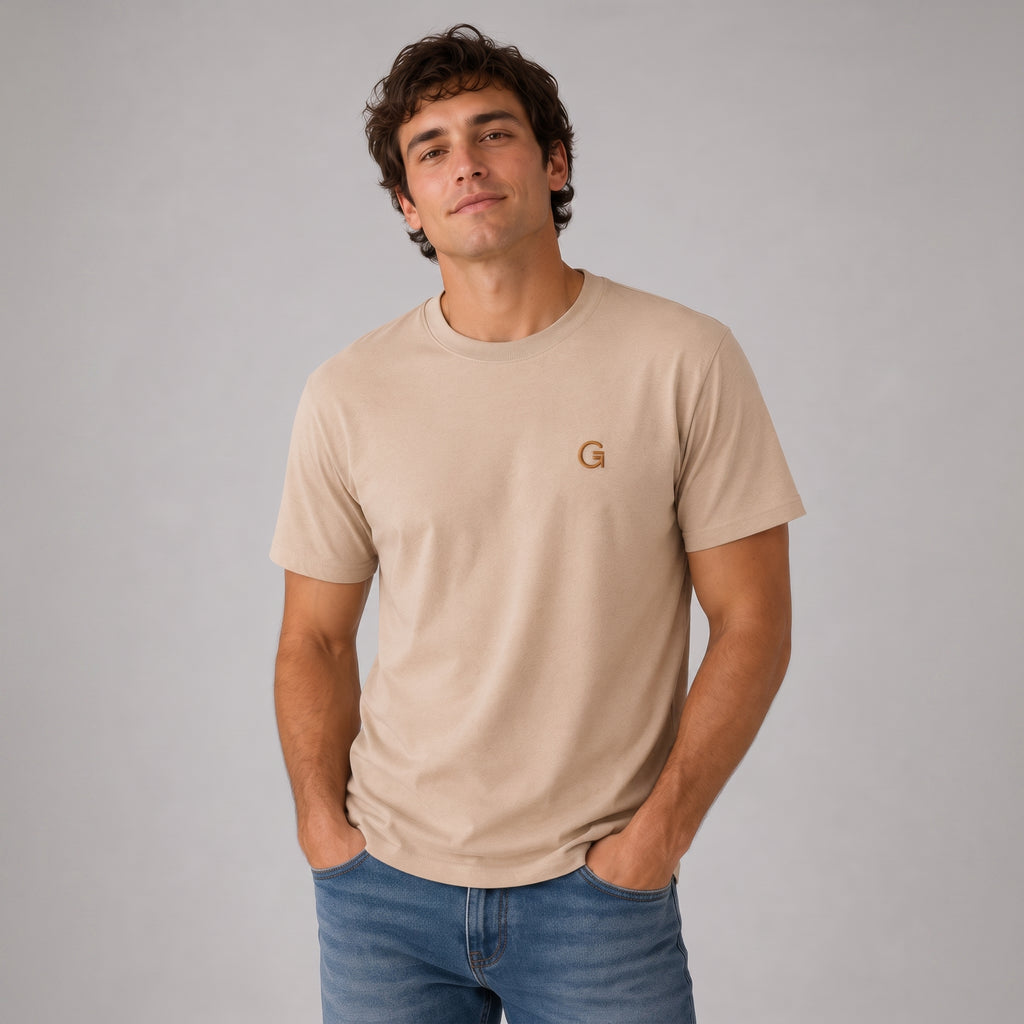 A man modelling a sandstone tee with a russet embroidered ‘Clobber & Gear’ logo on the left chest, paired against a light grey studio background.