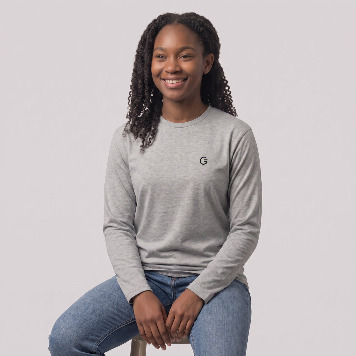 A woman modelling a heather grey long sleeved t-shirt with a black embroidered ‘Clobber & Gear’ logo on the left chest, paired against a light grey studio background.