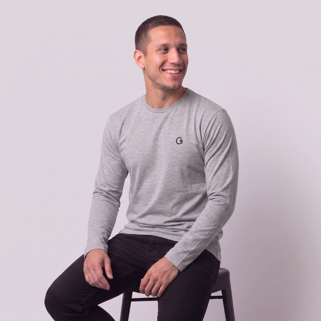 A man modelling a heather grey long sleeved t-shirt with a black embroidered ‘Clobber & Gear’ logo on the left chest, paired against a light grey studio background.