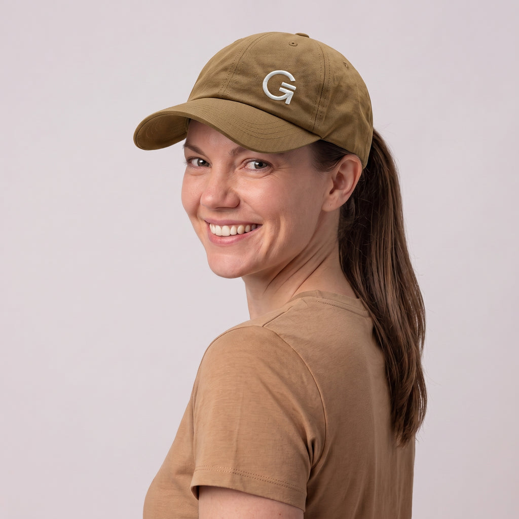A woman wearing a tan baseball cap with an offset white embroidered "Clobber and Gear" logo set against a light grey studio background.