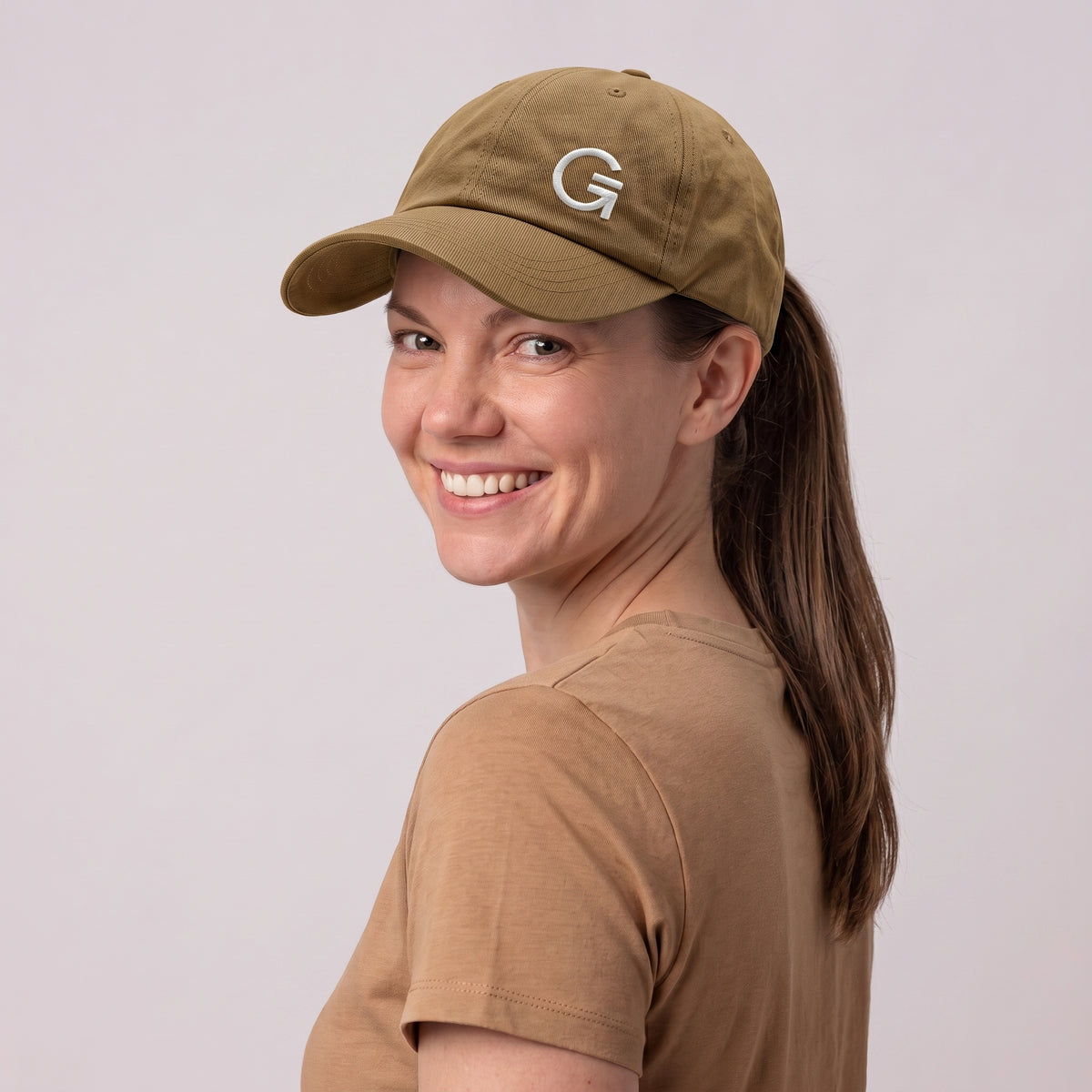 A woman wearing a tan baseball cap with an offset white embroidered "Clobber and Gear" logo set against a light grey studio background.