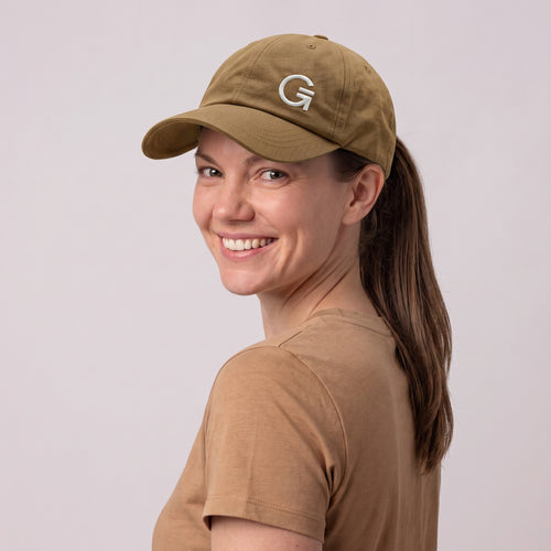 A woman wearing a tan baseball cap with an offset white embroidered "Clobber and Gear" logo set against a light grey studio background.