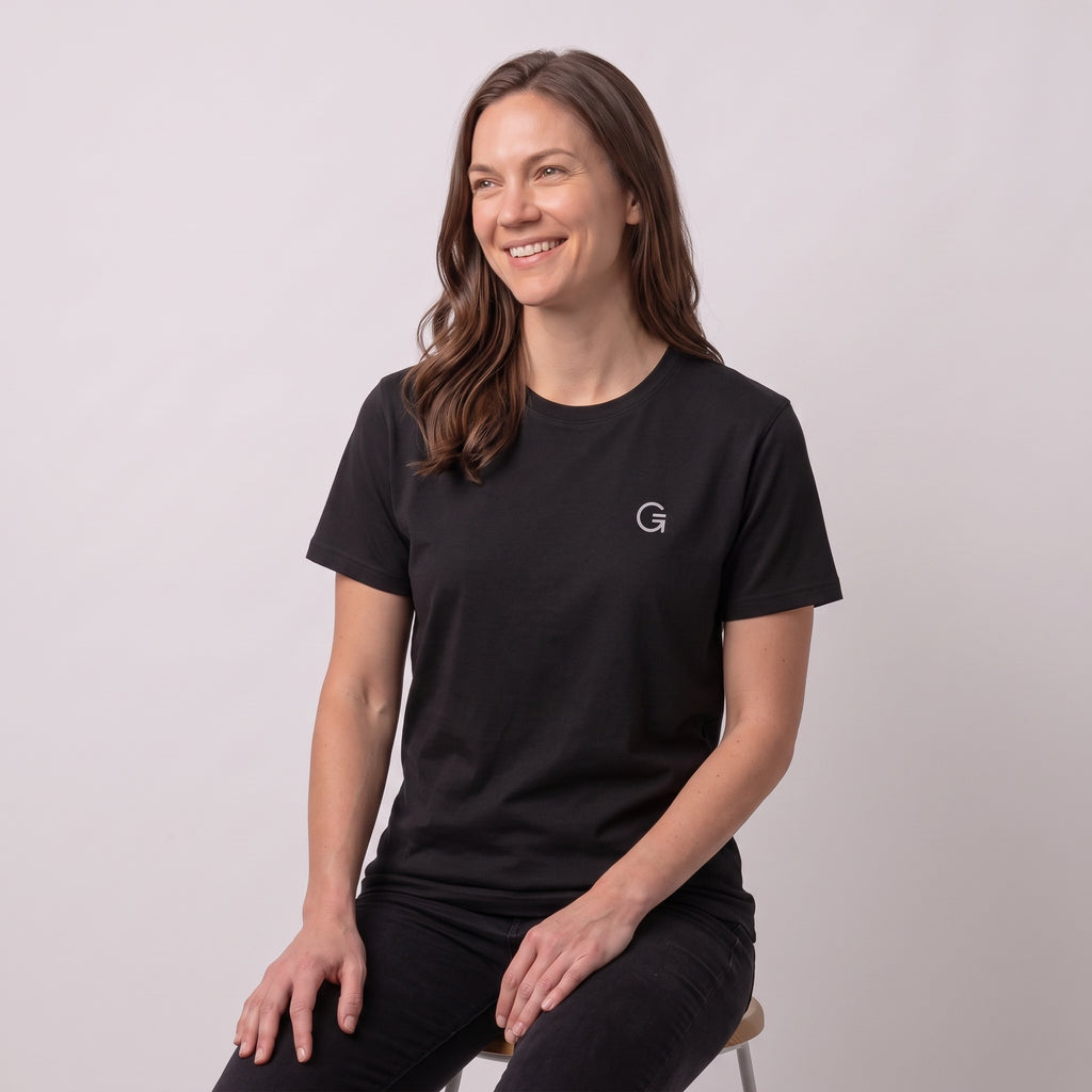 A woman modelling a black tee with a white embroidered ‘Clobber & Gear’ logo on the left chest, paired against a light grey studio background.