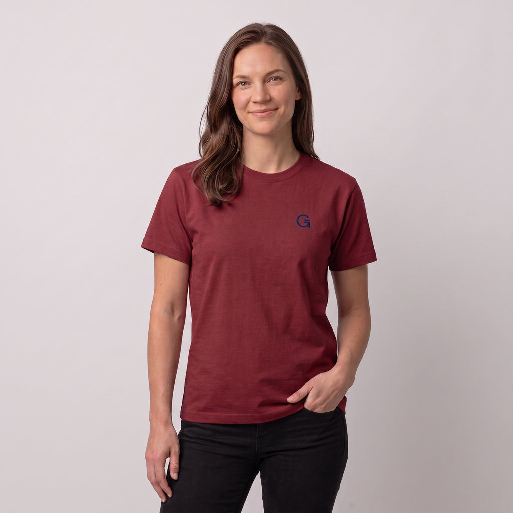 A woman modelling a brick red tee with a navy embroidered ‘Clobber & Gear’ logo on the left chest, paired against a light grey studio background.