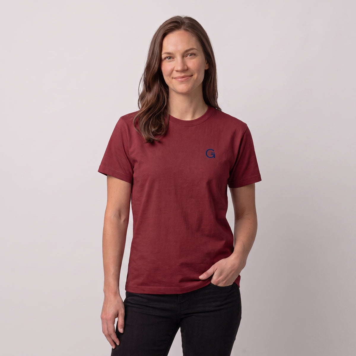 A woman modelling a brick red tee with a navy embroidered ‘Clobber & Gear’ logo on the left chest, paired against a light grey studio background.