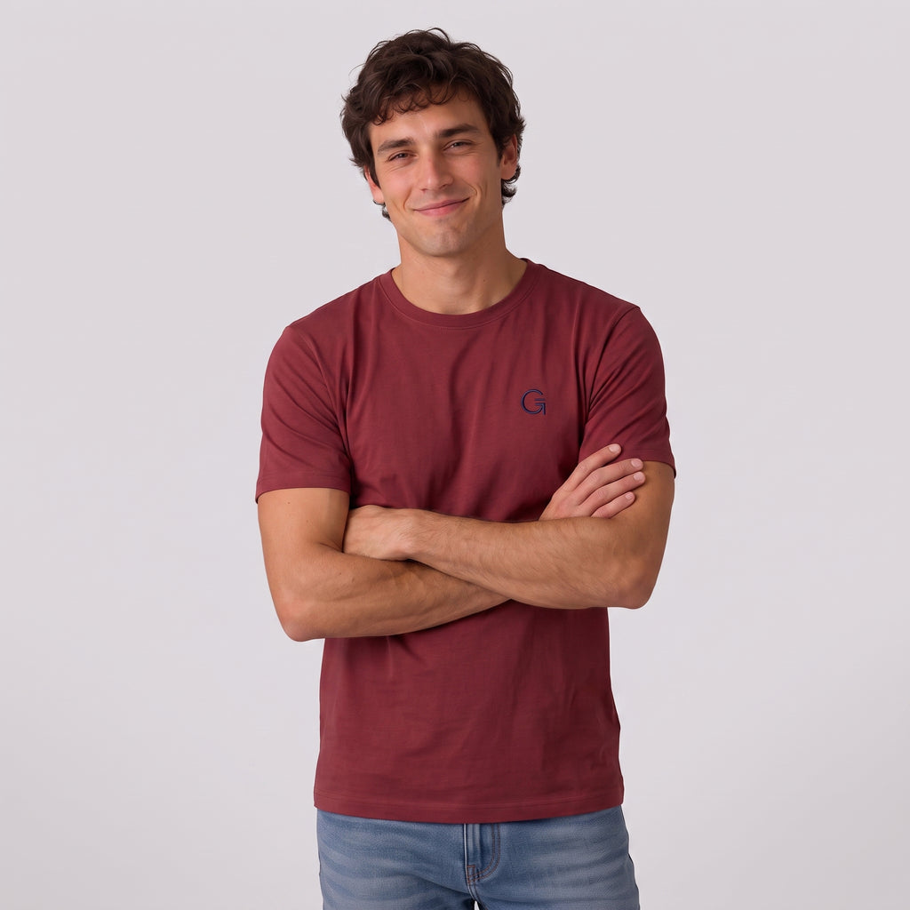 A man modelling a brick red tee with a navy embroidered ‘Clobber & Gear’ logo on the left chest, paired against a light grey studio background.