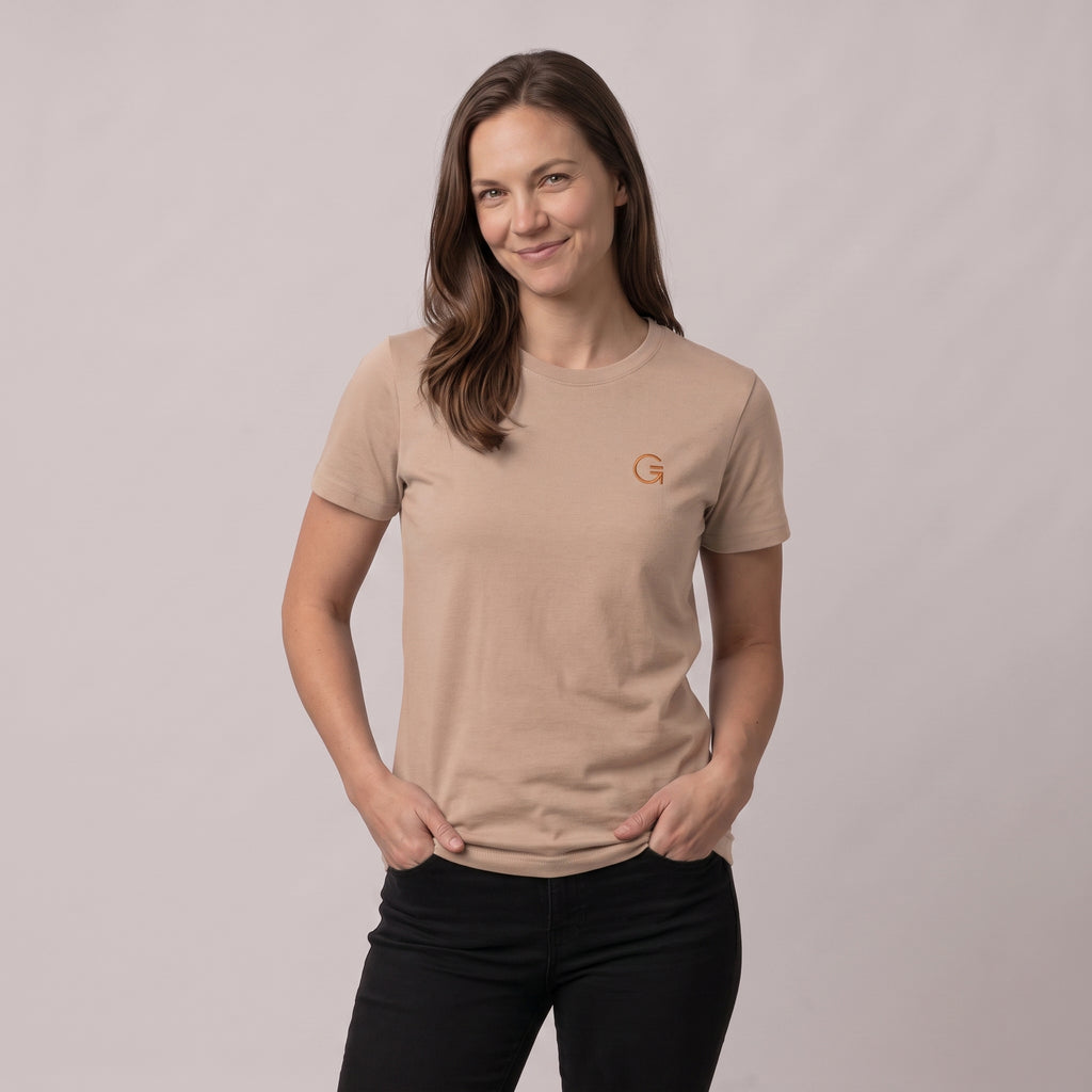 A woman modelling a latte colour tee with a burnt orange embroidered ‘Clobber & Gear’ logo on the left chest, paired against a light grey studio background.
