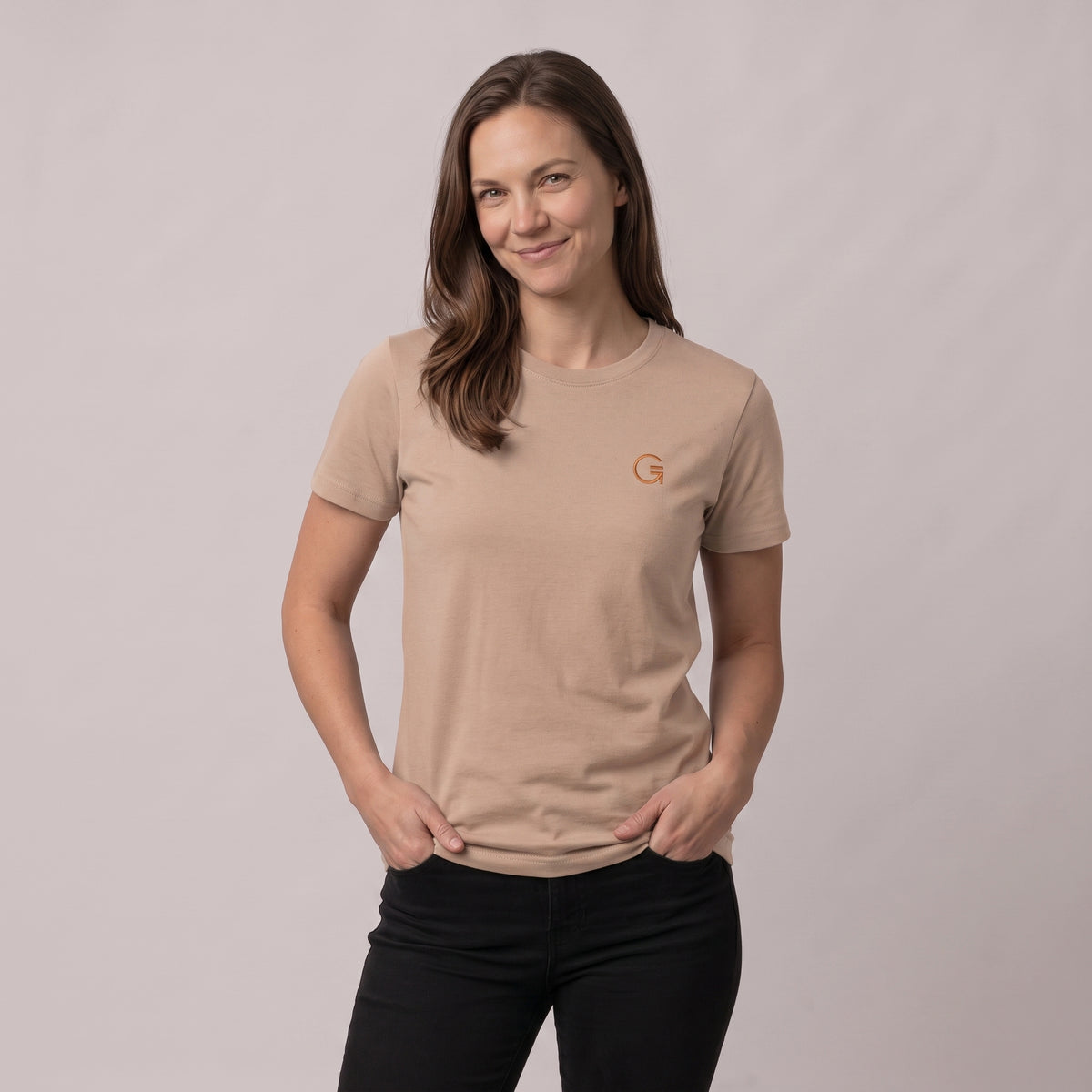A woman modelling a latte colour tee with a burnt orange embroidered ‘Clobber & Gear’ logo on the left chest, paired against a light grey studio background.