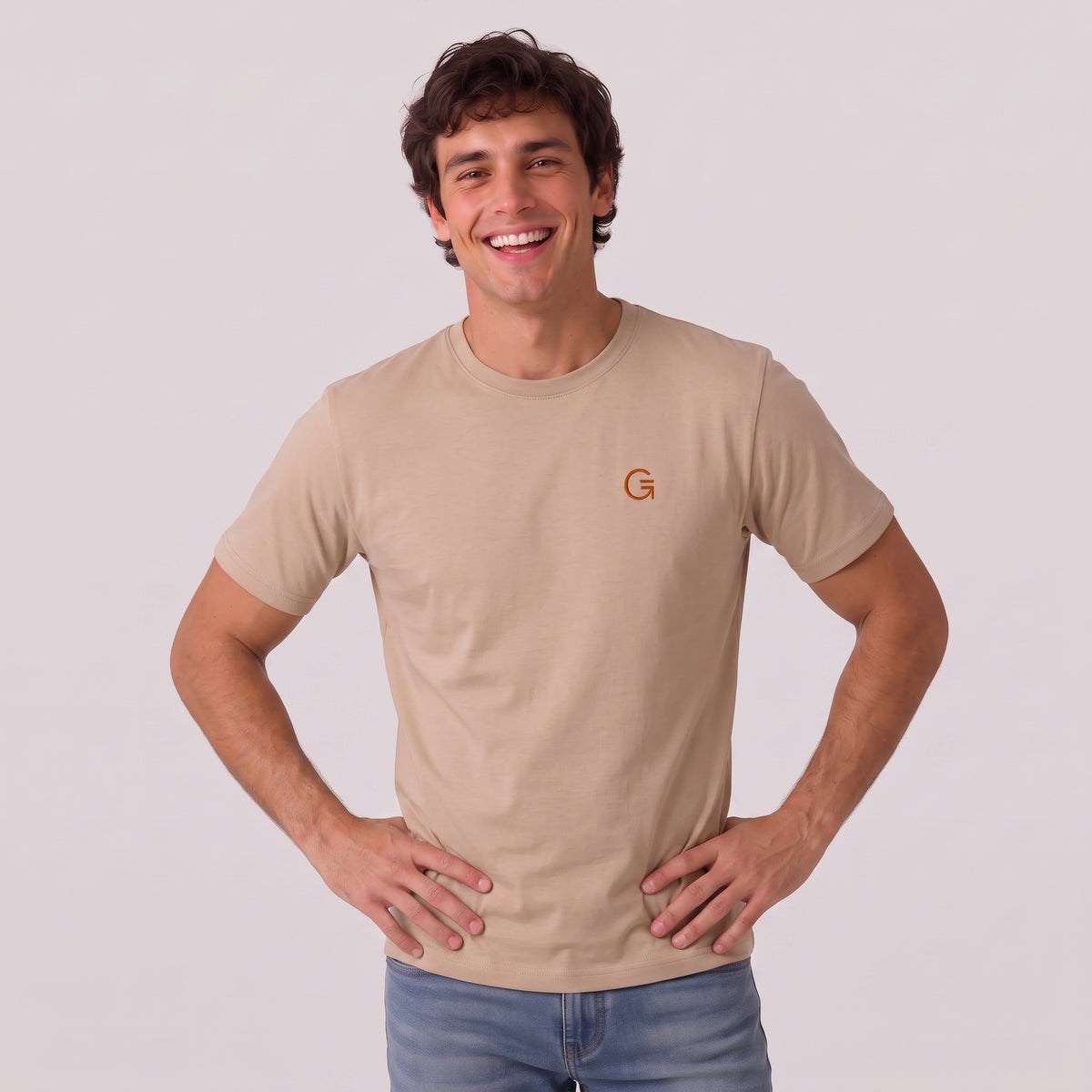 A man modelling a latte colour tee with a burnt orange embroidered ‘Clobber & Gear’ logo on the left chest, paired against a light grey studio background.